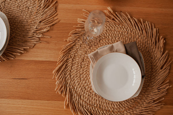 Minimalizmde çevre dostu masa, samandan yapılmış doğal yuvarlak bir peçete üzerinde beyaz bir tabak, ahşap bir masanın üstünde pamuk peçeteler. - Fotoğraf, Görsel