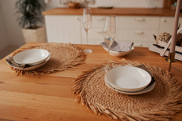 tavola eco-friendly in minimalismo, piatti bianchi profondi su un tovagliolo rotondo naturale di fieno, con tovaglioli di cotone, bicchieri di madreperla per champagne, su un tavolo di legno in una cucina leggera - Foto, immagini