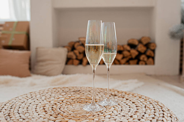 two mother-of-pearl glasses with champagne, bubbles and foam, on a wicker table, against the background of a bright interior with logs - Zdjęcie, obraz