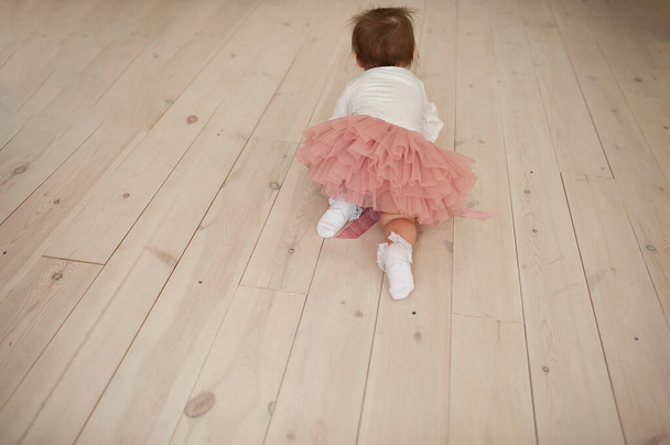 baby girl in a dusty pink tulle skirt, white lace socks crawls on a light wooden floor, rear view - Photo, Image