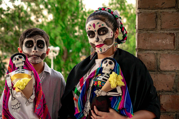 Δύο έφηβοι που φορούν παραδοσιακή Ημέρα των Νεκρών μακιγιάζ, κρατώντας παραδοσιακά μεξικάνικα κρανία με φόντο ένα δέντρο. - Φωτογραφία, εικόνα