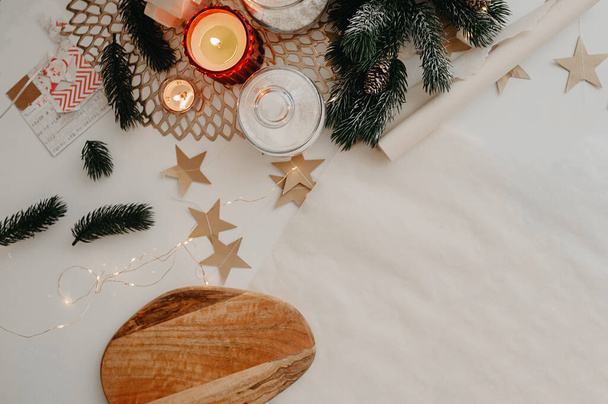 Kerstversiering, een rol bakpapier, brandende witte en rode kaarsen, houten snijplank, kerstboomtakjes en papieren sterren, liggen op een wit tafelblad - Foto, afbeelding