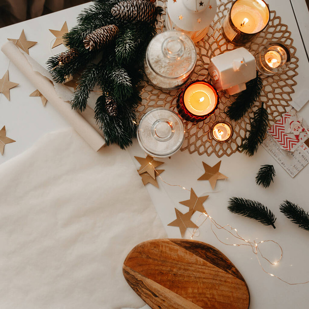 Weihnachtsdekoration, eine Rolle Backpapier, brennende weiße und rote Kerzen, Holzschneidebrett, Weihnachtsbaumzweige und Papiersterne, liegen auf einem weißen Tisch, quadratische Ernte - Foto, Bild
