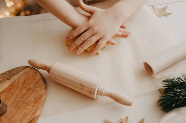 dětské ruce válejí těsto s malou dřevěnou válečkovou jehlicí na pečicím papíru na bílém stole - Fotografie, Obrázek