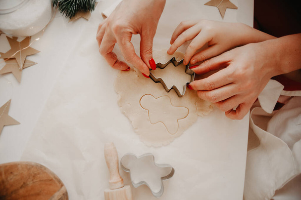 hands of mom and baby cut out cookies from the dough using metal molds, on baking paper, on a white table with decor from paper stars - Φωτογραφία, εικόνα