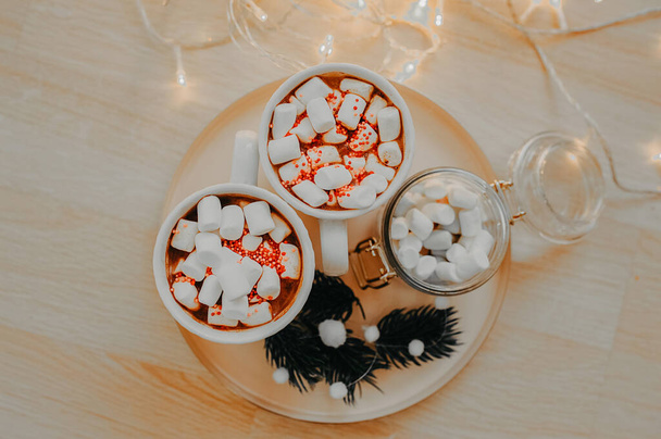 two white cups with a hot drink and white marshmallows, on a golden tray with a glass jar of white marshmallows, sprigs of a Christmas tree, on a wooden floor in a large number of lights from a garland - Photo, Image