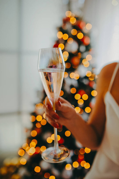 ragazza in abiti bianchi con le mani nude tiene un bel vetro di madreperla con champagne su uno sfondo sfocato albero di Natale - Foto, immagini
