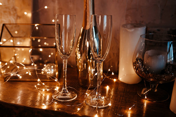 δύο άδεια ποτήρια και ένα χρυσό μπουκάλι σε ένα ξύλινο τραπέζι σε γιρλάντες και βολβούς στο σκοτάδι, τραπέζι για μια ρομαντική βραδιά για δύο εραστές - Φωτογραφία, εικόνα