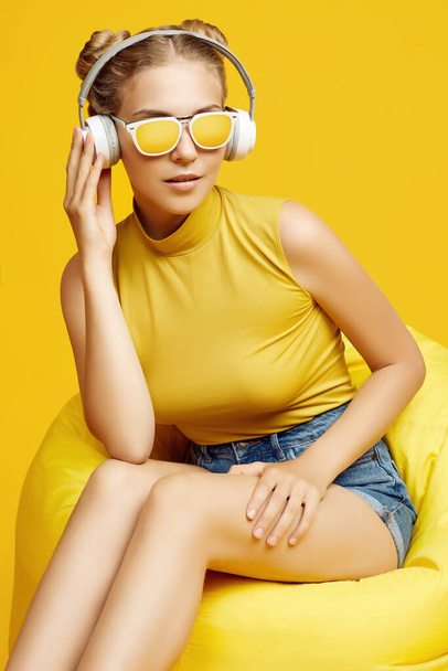 Porträt eines wunderschönen blonden Hipster-Mädchens mit Sonnenbrille, das in einem Sacksack mit Kopfhörern auf buntem gelben Hintergrund sitzt - Foto, Bild