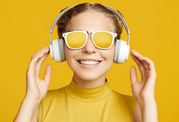 Portrait of gorgeous blonde hipster girl with curly hair wearing sunglasses enjoys the music in headphones on yellow background - Photo, image