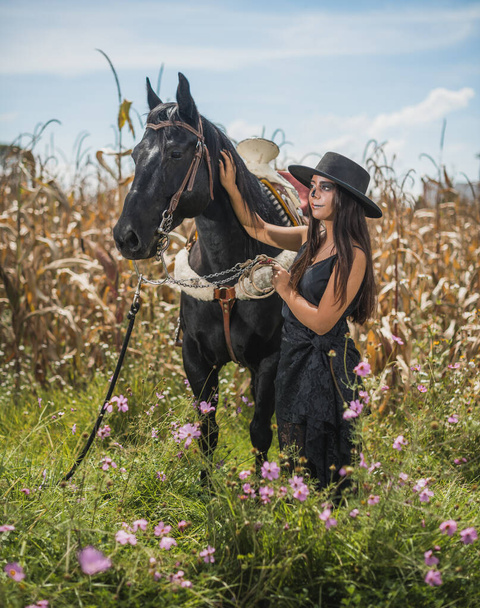 Girl disguised as Catrina (Mexican female character of death) dressed in black, in the middle of a corn field, with mirasoles flowers, stroking a horse 12 - Photo, Image