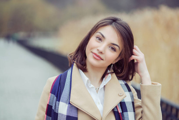 Young girl in coat looking away. Closeup portrait headshot of beautiful stylish smiling  fixing hair, scratching head girl student isolated cityscape park lake background.  Multicultural, mixt race - Foto, immagini