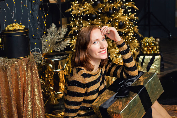 Eine junge Frau mit Weihnachtsgeschenkschachteln hält Geschenke vor dem Hintergrund eines Weihnachtsbaums mit Goldschmuck. Stilvolle Damenmode, modische Abendkleidung und Accessoires, Make-up. Frohes neues Jahr. - Foto, Bild