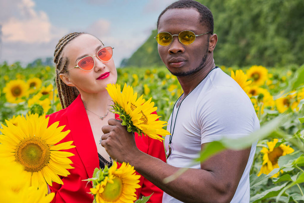 Ein junges verliebtes Paar, ein Mann und ein Mädchen, stehen inmitten eines Sonnenblumenfeldes und blicken in die Kamera - Foto, Bild