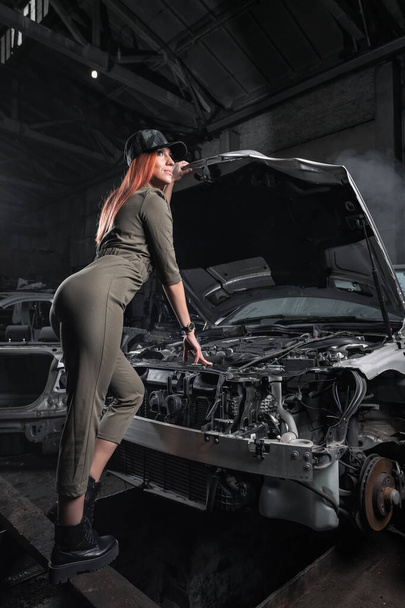 Model in stylish clothes standing against on open hood in disassembled car in the garage. Design Car workshop art concept. Woman repairing an old car in a workshop - Foto, Imagem