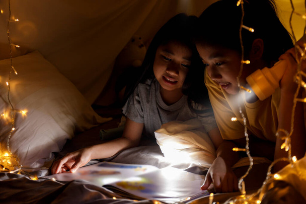 Семейная концепция. старшая сестра и сестра читают вместе книгу с фонариком перед сном. Сестра вместе читала сказки в палатке с простынями. Сосредоточься на сестре. С эффектом зерна пленки - Фото, изображение