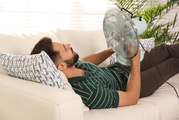 Man with fan lying on sofa home. Summer heat - Foto, Imagen