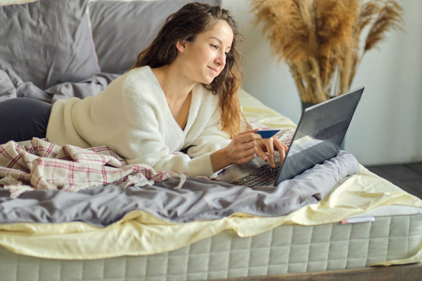 Frau kauft online mit Kreditkarte und Laptop ein, während sie zu Hause im Bett sitzt - Foto, Bild