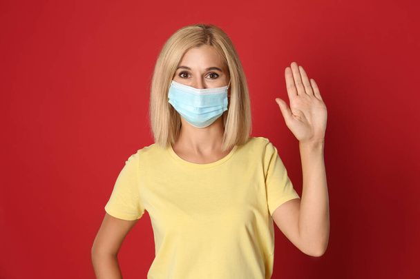 Woman in protective mask showing hello gesture on red background. Keeping social distance during coronavirus pandemic - Photo, Image