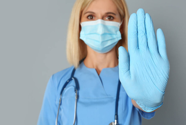Doctor in protective mask showing stop gesture on grey background. Prevent spreading of coronavirus - Фото, зображення