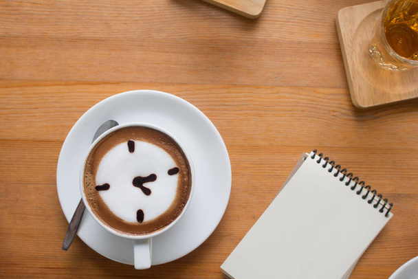 Lähikuva A Cup kuuma latte art kahvi piirretty oclock liiketoiminnan käsite puinen pöytä muistiinpanokirja. - Valokuva, kuva