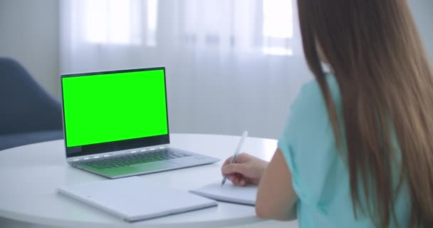 Vrouw zit aan een bureau in de slaapkamer, ze kijkt naar laptop groen scherm en praat met iemand via internet video communicatie, soms notities maken in notebook. Close-up - Video