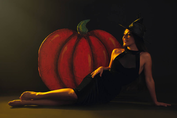 Young woman wearing witch costume near decorative pumpkin on black background. Halloween party - Φωτογραφία, εικόνα
