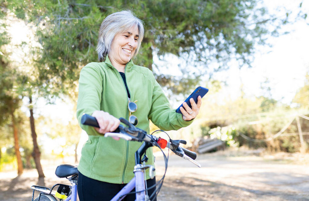 ηλικιωμένη γυναίκα στο πάρκο χρησιμοποιώντας smartphone και ποδήλατο - Φωτογραφία, εικόνα