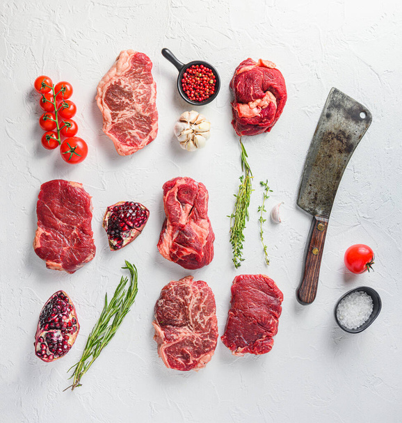 Rraw beef steaks set top blade, romp, chuck eye roll met kruiden granaatappel en slagersmes voor het hakken over witte beton achtergrond, bovenaanzicht - Foto, afbeelding