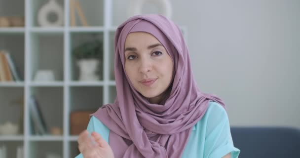Une musulmane dans un hijab s'assoit à une table et regarde dans la caméra en agitant la main et en parlant. vlog ou vidéo appeler une femme dans un hijab. coup de feu de la tête d'une femme arabe en conversation - Séquence, vidéo