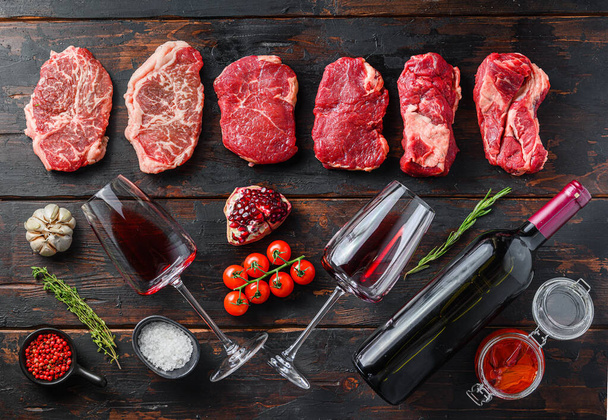 Organikus alternatív marhahús darabok felső penge steak, Chuck roll, hátsósteak nyers marhahús fűszerezéssel, rozmaring és hentes bárd és vörösbor palack. Fa textúrájú háttér. Top view panoráma nagy méret - Fotó, kép
