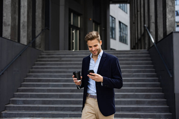 Handsome office worker holding smartphone and smiling. Happy young man using mobile phone apps, texting message, browsing internet, looking at smartphone. Coffee break near the business center - Foto, imagen