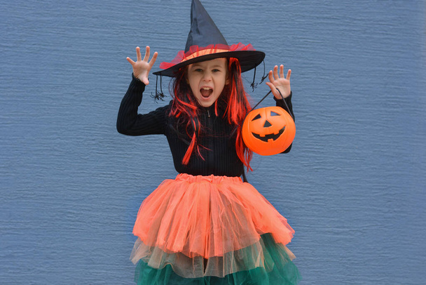 Doces ou truques de Halloween. Rapariga sorridente com um chapéu de bruxa contra uma parede azul. Doce ou travessura! Espaço de cópia para texto - Foto, Imagem