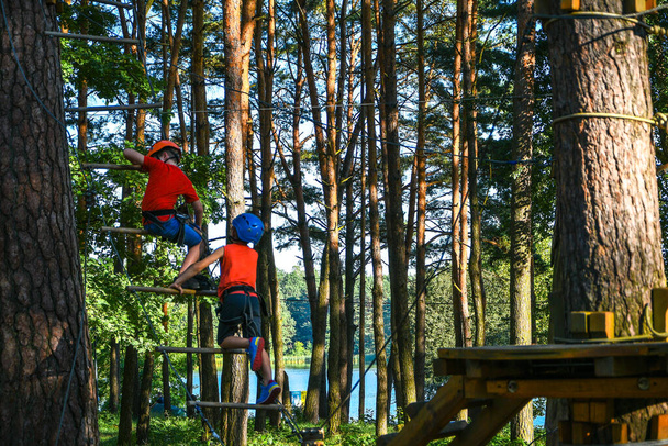 Adventure climbing high wire park. Close-up. Young boy in helmet having fun and playing at adventure park, holding ropes and climbing wooden stairs. active lifestyle concept. Summer leisure activities - Photo, Image