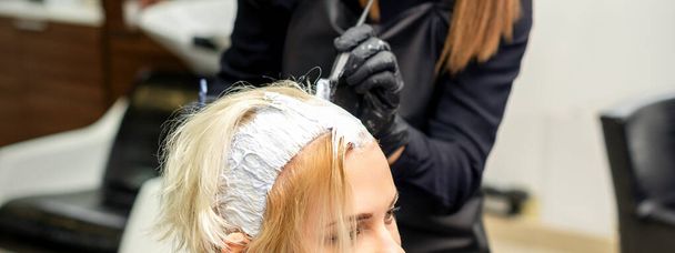 Женский стилист наносит белый краситель на волосы молодой клиентки в парикмахерской - Фото, изображение
