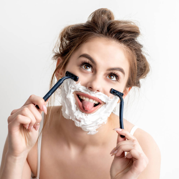Beautiful young caucasian woman shaving her face by two razors with tongue out on white background. - Photo, Image