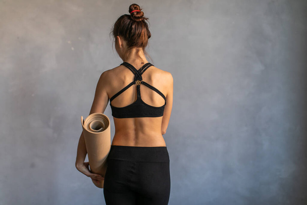 Woman standing at work out or yoga class with rolled fitness mat in her hads turned her back to the camera. Copy space on dark background - Foto, Imagem