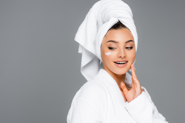 usmívající se žena s ručníkem na hlavě a kosmetický krém na tváři izolované na šedé - Fotografie, Obrázek