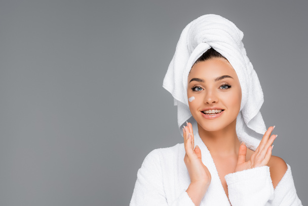 smiling woman with towel on head and cosmetic cream on face isolated on grey - Foto, afbeelding