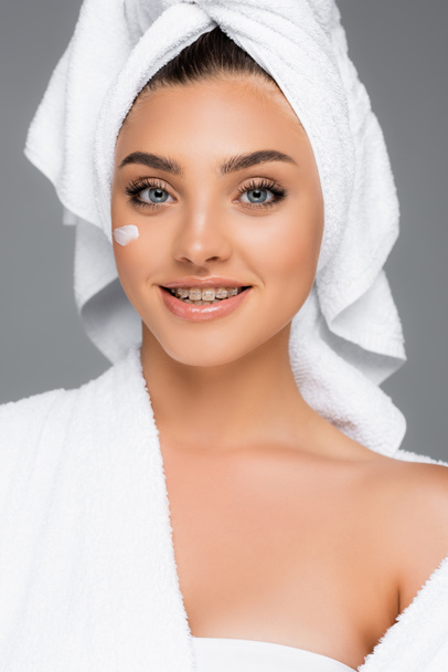 donna sorridente con asciugamano sulla testa e crema cosmetica sul viso isolata sul grigio - Foto, immagini
