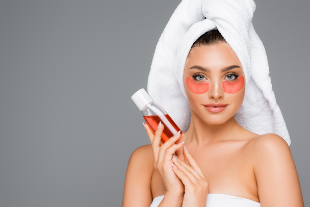 kobieta z ręcznikiem na głowie i przepaskach na oczy trzymając balsam izolowany na szarości - Zdjęcie, obraz