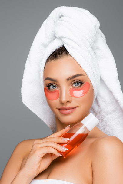 woman with towel on head and eye patches holding lotion isolated on grey - Foto, Bild