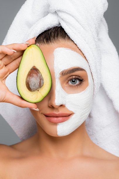 woman with towel on head and clay mask on face holding avocado isolated on grey - Foto, Imagem