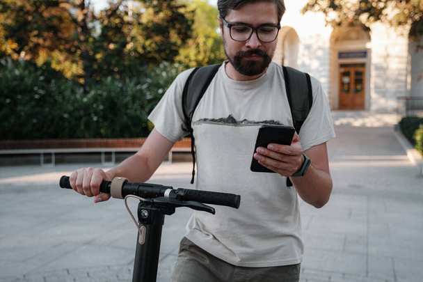 hipster man commuter with electric scooter outdoors in city, using smartphone. Young millennial guy using cellphone rental of eco transportation in summer - Foto, Imagen