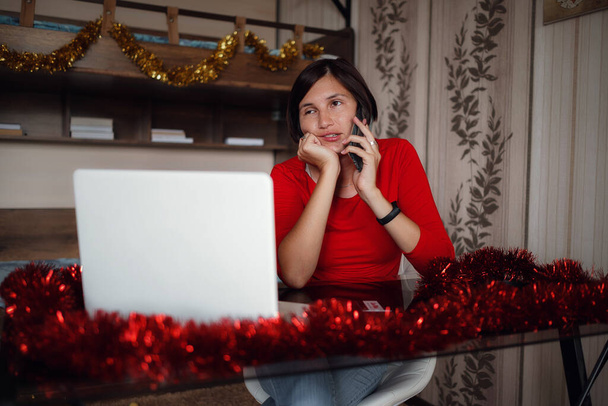 Concepto de Navidad compras en línea, la compra de regalos, felicitaciones a través de Internet. Linda dama se sienta con el ordenador portátil, mira a la pantalla. Cajas de regalo cercanas. blanco y rojo - Foto, Imagen