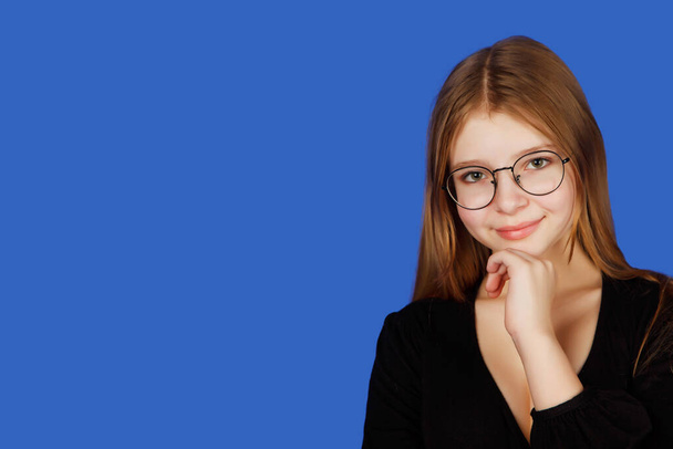 Emotional thirteen-year-old cute teenage girl in glasses thinking and look at camera flirting, isolated against blue Royal background. Caucasian child in black clothes shows love emotions. Copy space - Photo, image