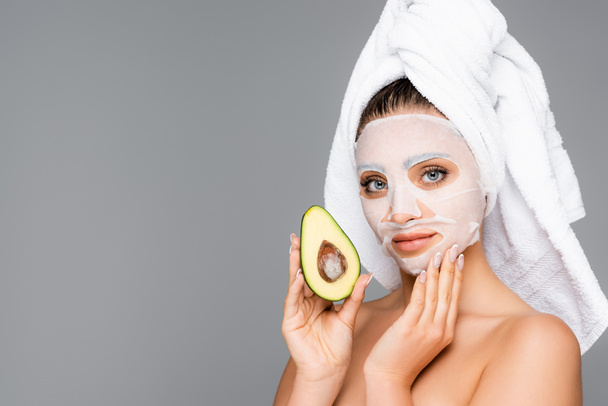 женщина с полотенцем на голове и маска лист на лице держа авокадо изолированы на серый - Фото, изображение