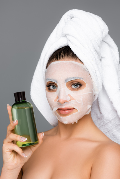 donna con asciugamano sulla testa e lenzuolo maschera sul viso bottiglia tenuta isolata su grigio - Foto, immagini
