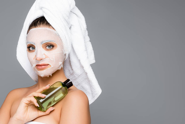 kobieta z ręcznikiem na głowie i arkusz maski na twarzy gospodarstwa butelki izolowane na szaro - Zdjęcie, obraz