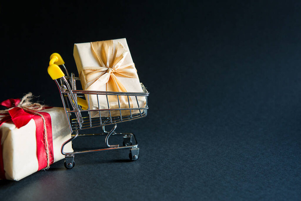 Box with a gift in Christmas packaging in a shopping cart on a black background. Black Friday, buying gifts for the new year. Space for text - 写真・画像